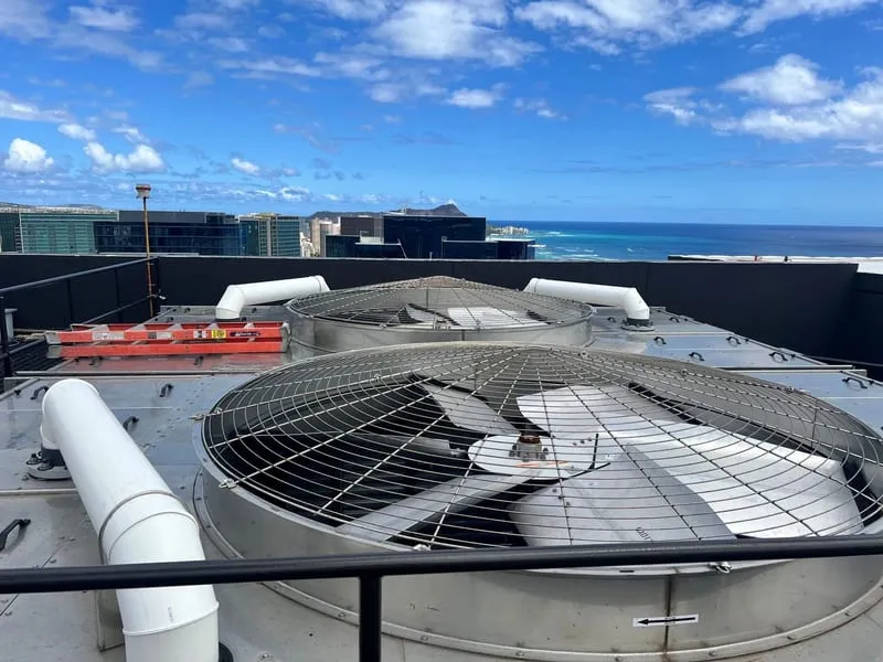 Two rooftop cooling tower fans, with a city skyline and open ocean beyond.