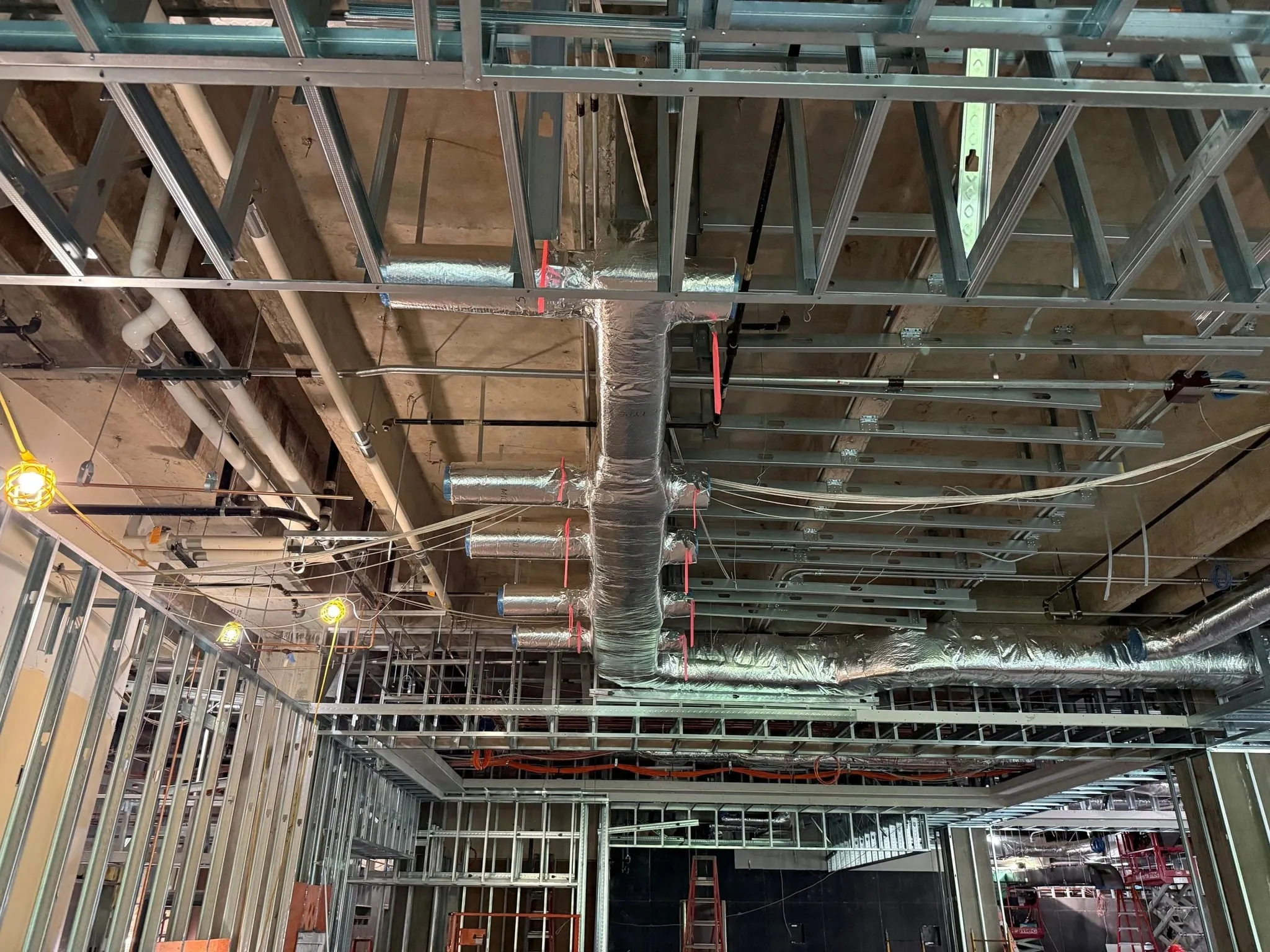 Overhead ductwork, flexible runs and unistrut trapeze above metal stud framing, before the ceiling is closed.
