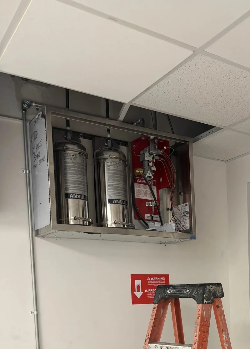 An opened kitchen fire suppression cabinet above a ceiling, showing two cylinders and the actuator.